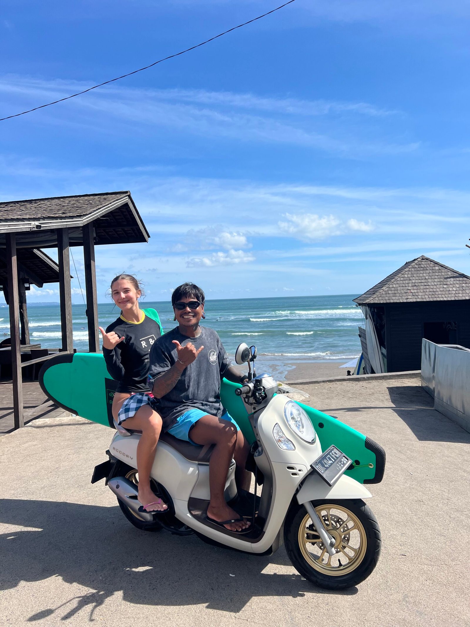 Made Moks and a guest riding a scooter with a surfboard